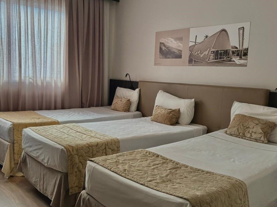 Quarto de hotel com três camas arrumadas, paredes bege e cortinas na janela.