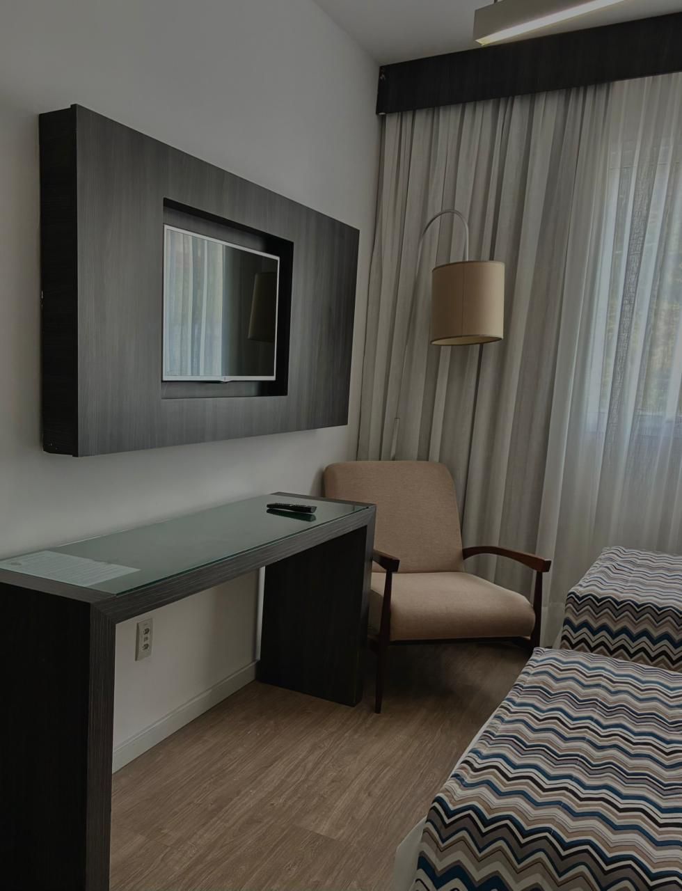 Quarto de hotel pequeno com escrivaninha, poltrona, abajur de chão, cortina e colcha estampada.
