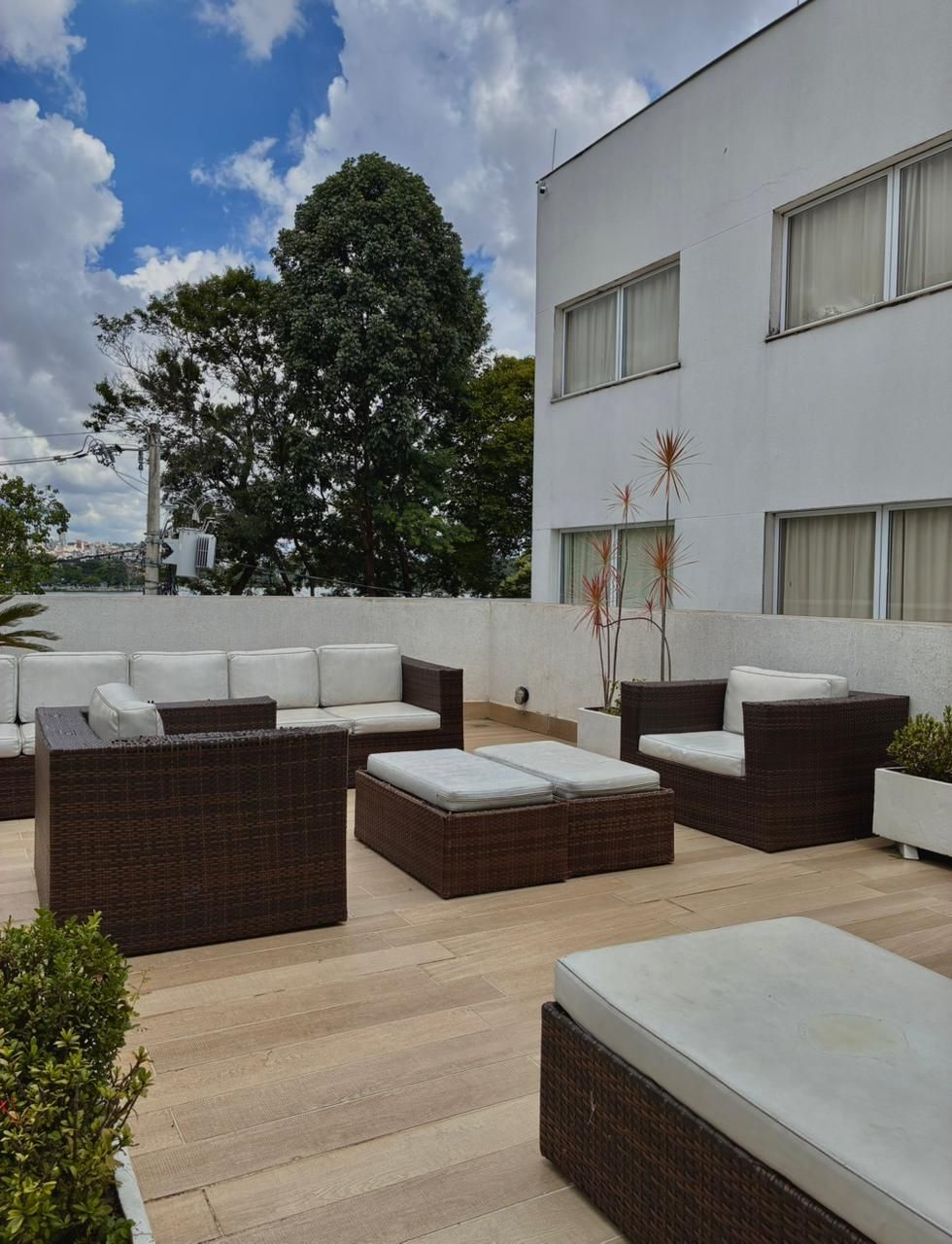 Terraço externo na cobertura com sofás de vime, almofadas brancas e vista para um prédio branco e árvores.