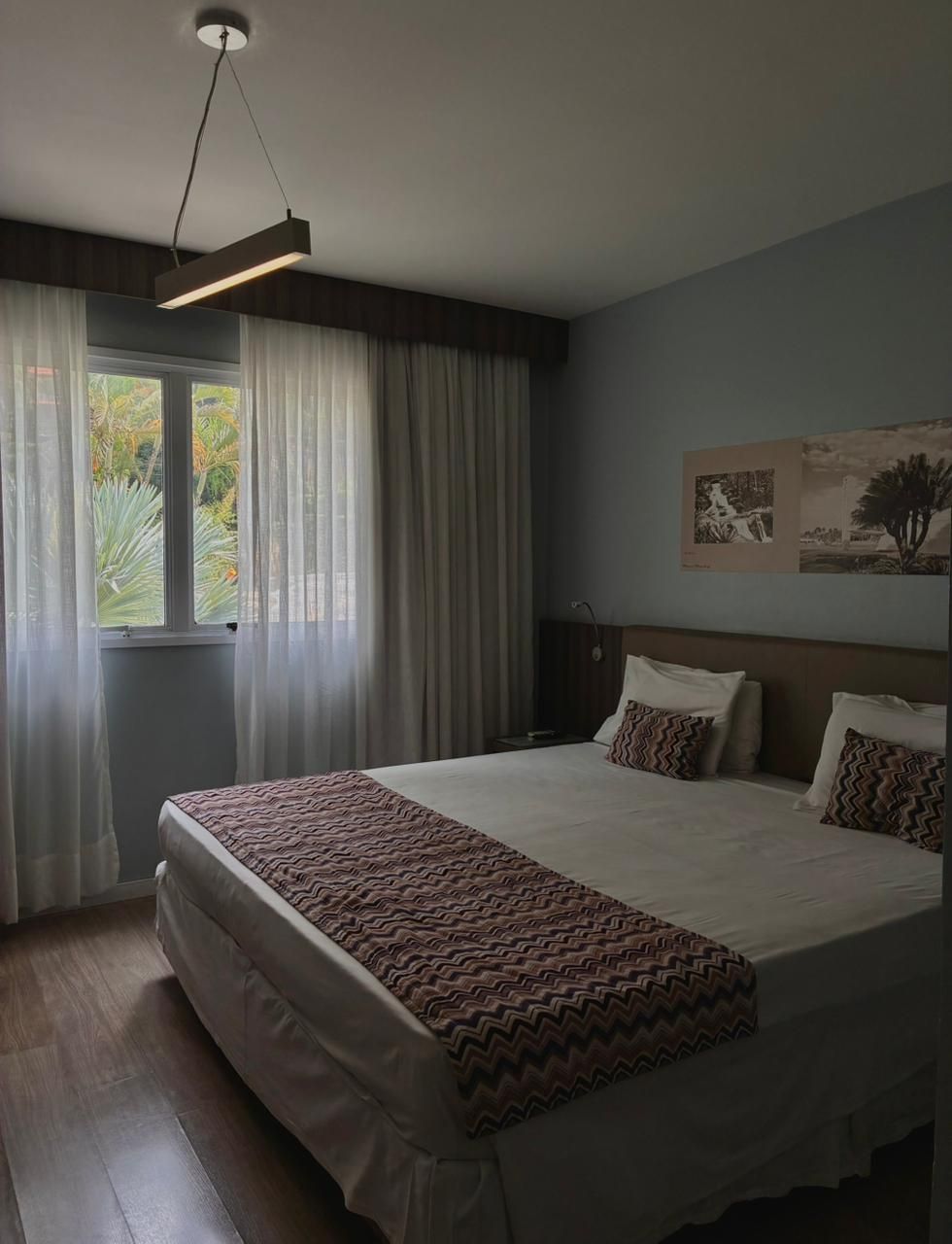 Quarto aconchegante com uma cama grande, cortinas cinza, iluminação suave e obras de arte na parede junto à janela.