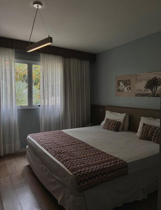 Quarto aconchegante com uma cama grande, cortinas cinza, iluminação suave e obras de arte na parede junto à janela.
