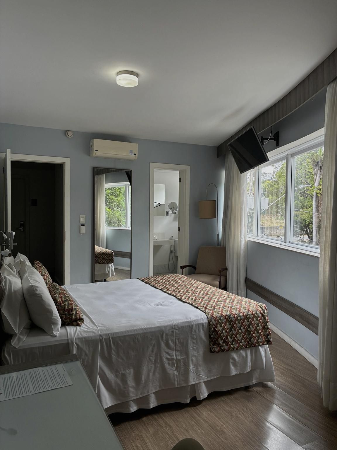 Quarto luminoso com cama, espelho de parede, janelas e uma pequena escrivaninha perto de uma porta de vidro deslizante.