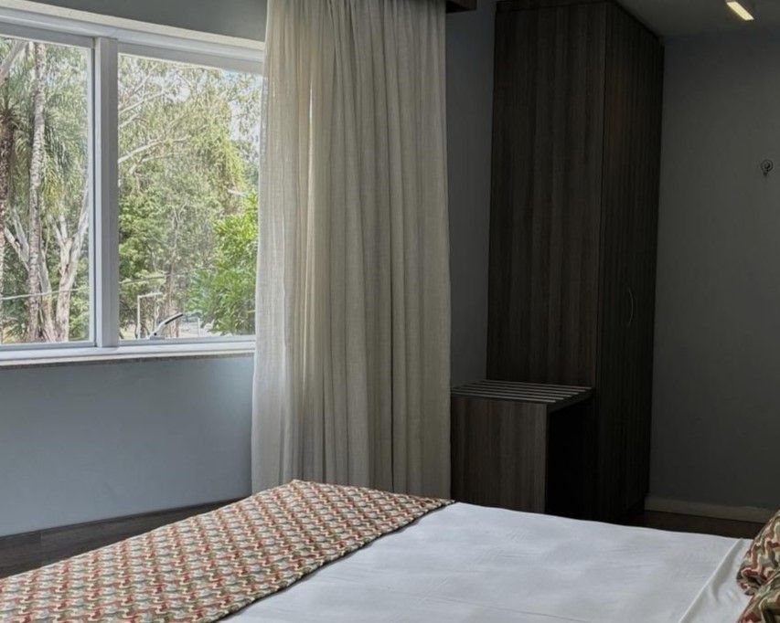 Quarto com cama, janela grande, cortinas brancas e um nicho escuro com guarda-roupa.