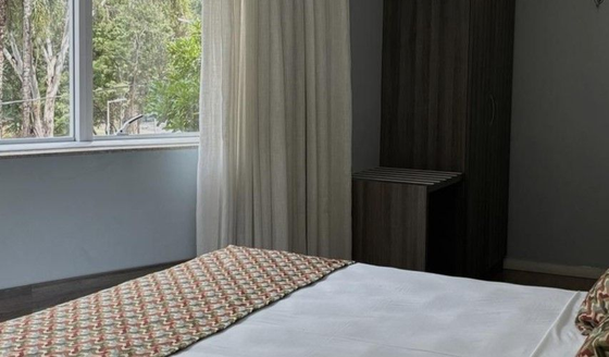 Uma cama arrumada junto à janela, com cortinas translúcidas, num quarto com iluminação suave.