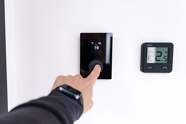 Hand pressing a black wall thermostat next to a digital temperature display on a white wall