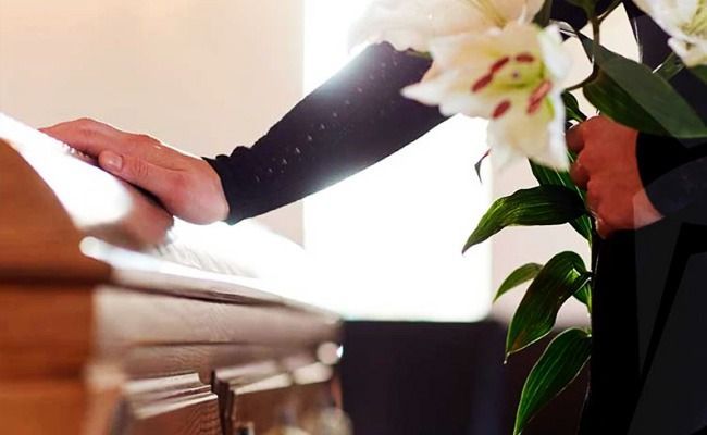 Una mano tocando un ataúd de madera en un funeral, con una flor blanca en primer plano.