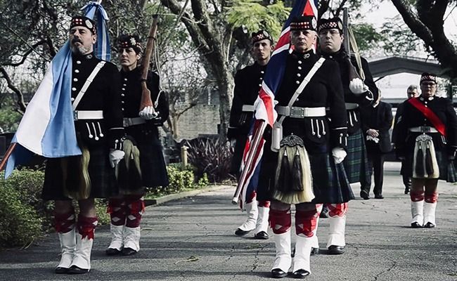 Gaiteros uniformados de negro y rojo marchan al aire libre en un desfile, uno de ellos portando una bandera azul y blanca.