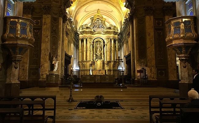 Gran interior de catedral con altar dorado, columnas ornamentadas y bancos en una luz cálida y tenue.