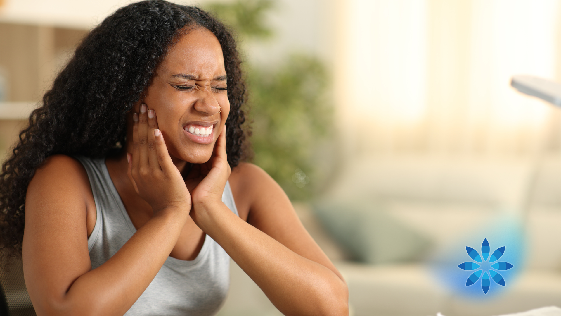 Person grimacing and holding jaw, seated on a couch in a bright room, suggesting tooth pain.