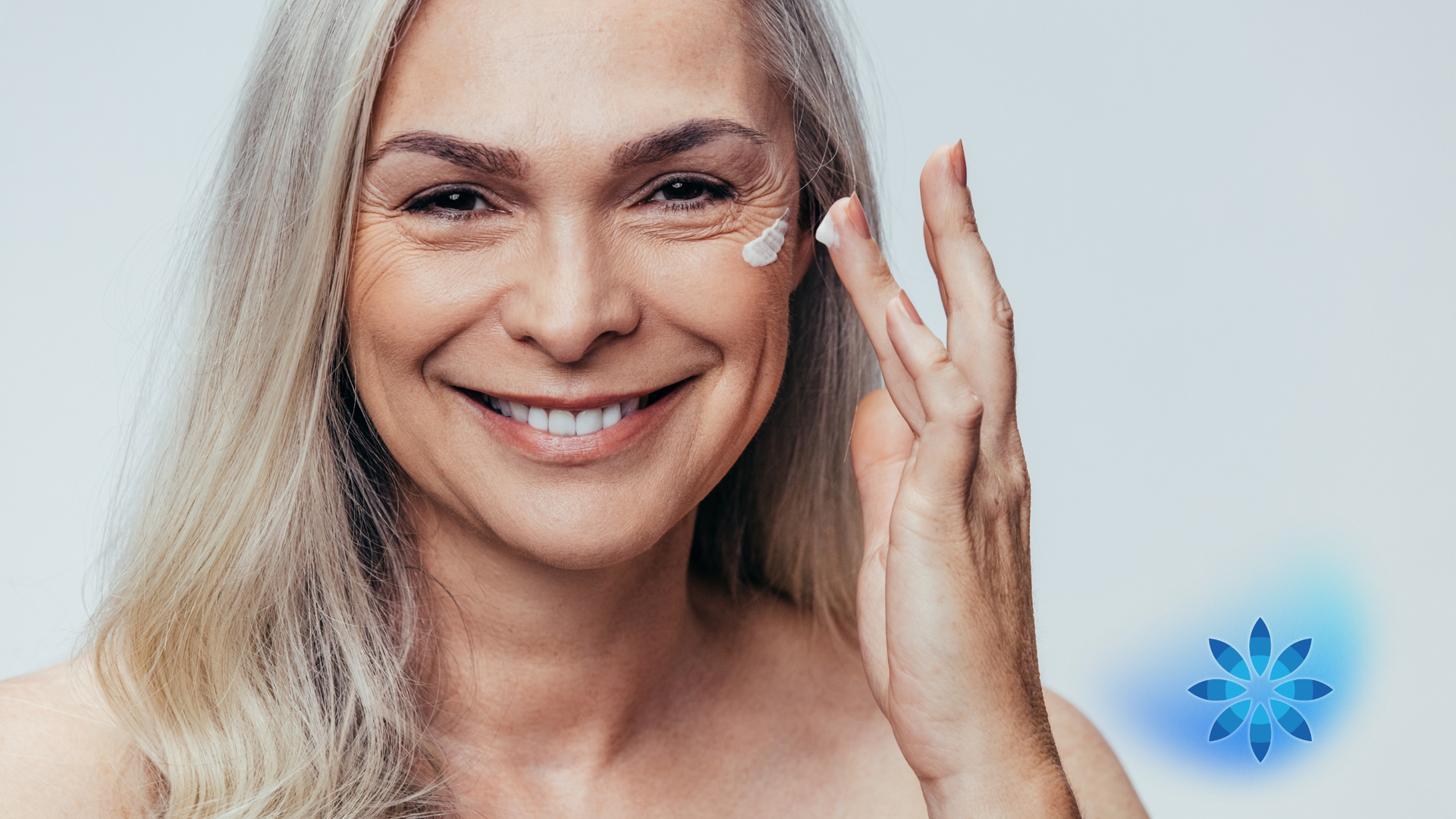 Woman applying cream under her eye, smiling. Gray hair, light background, blue snowflake graphic.