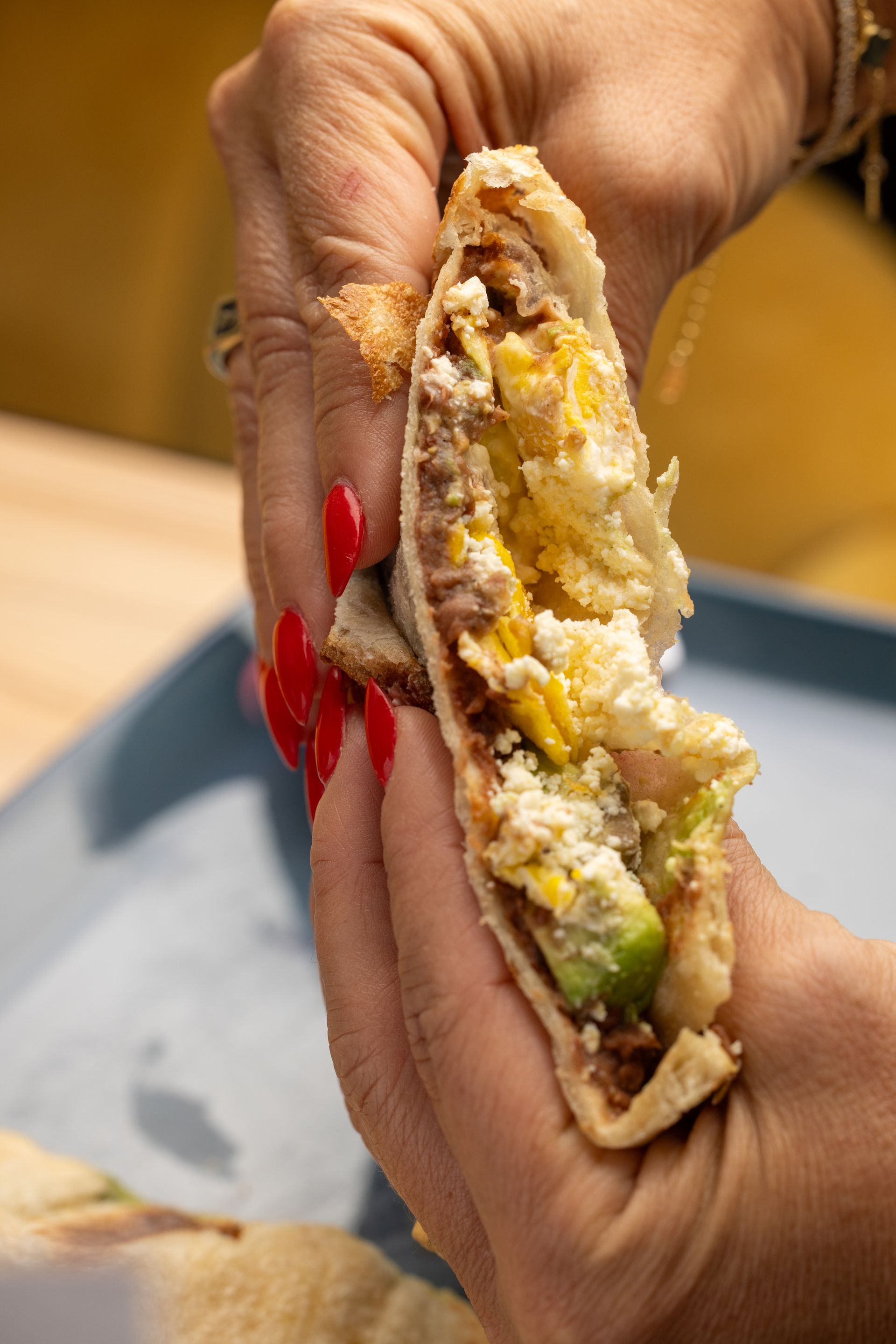 Person holding a pita sandwich, revealing fillings: avocado, cheese, and more. Red nails visible.