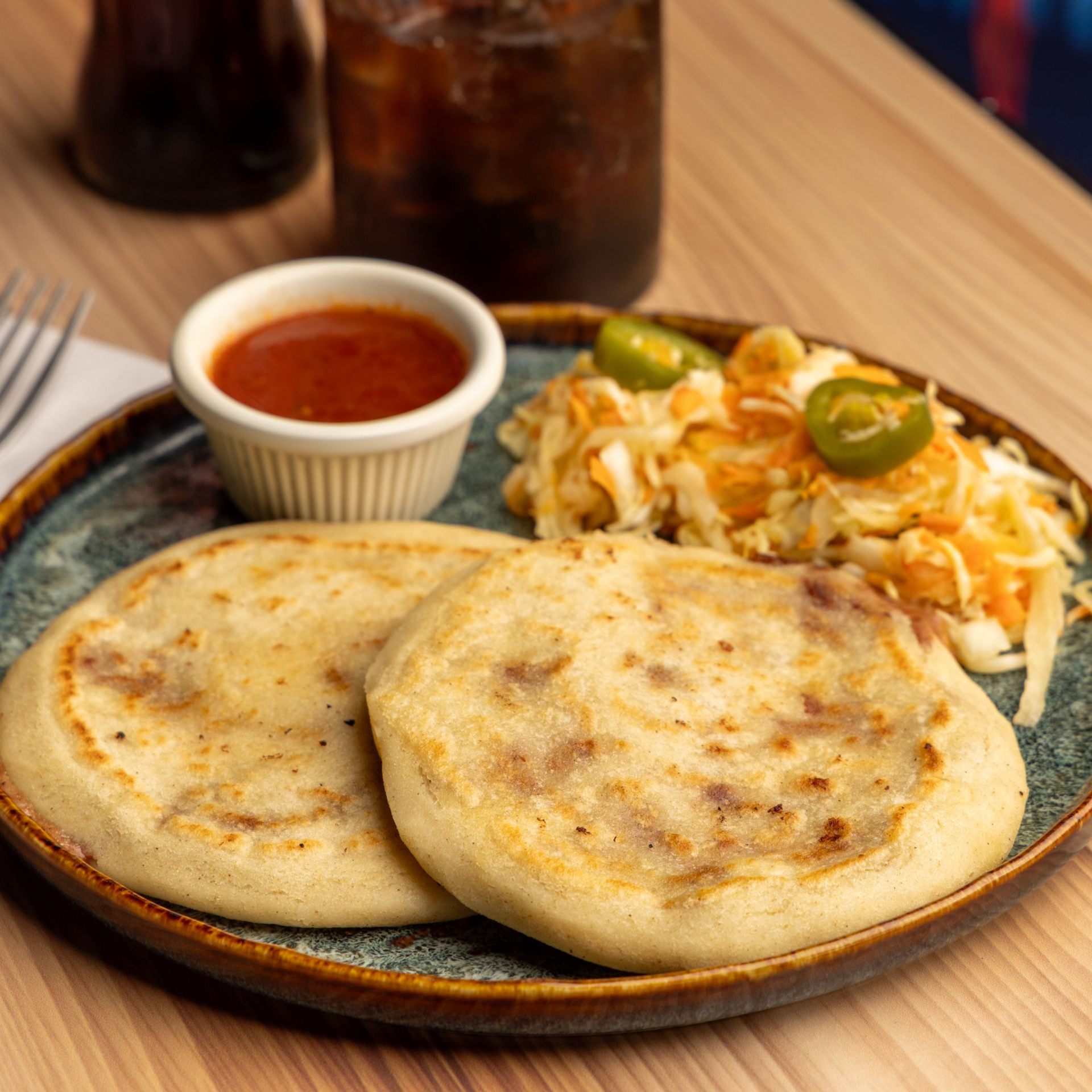 Two pupusas on a plate with slaw, salsa, and a soda.