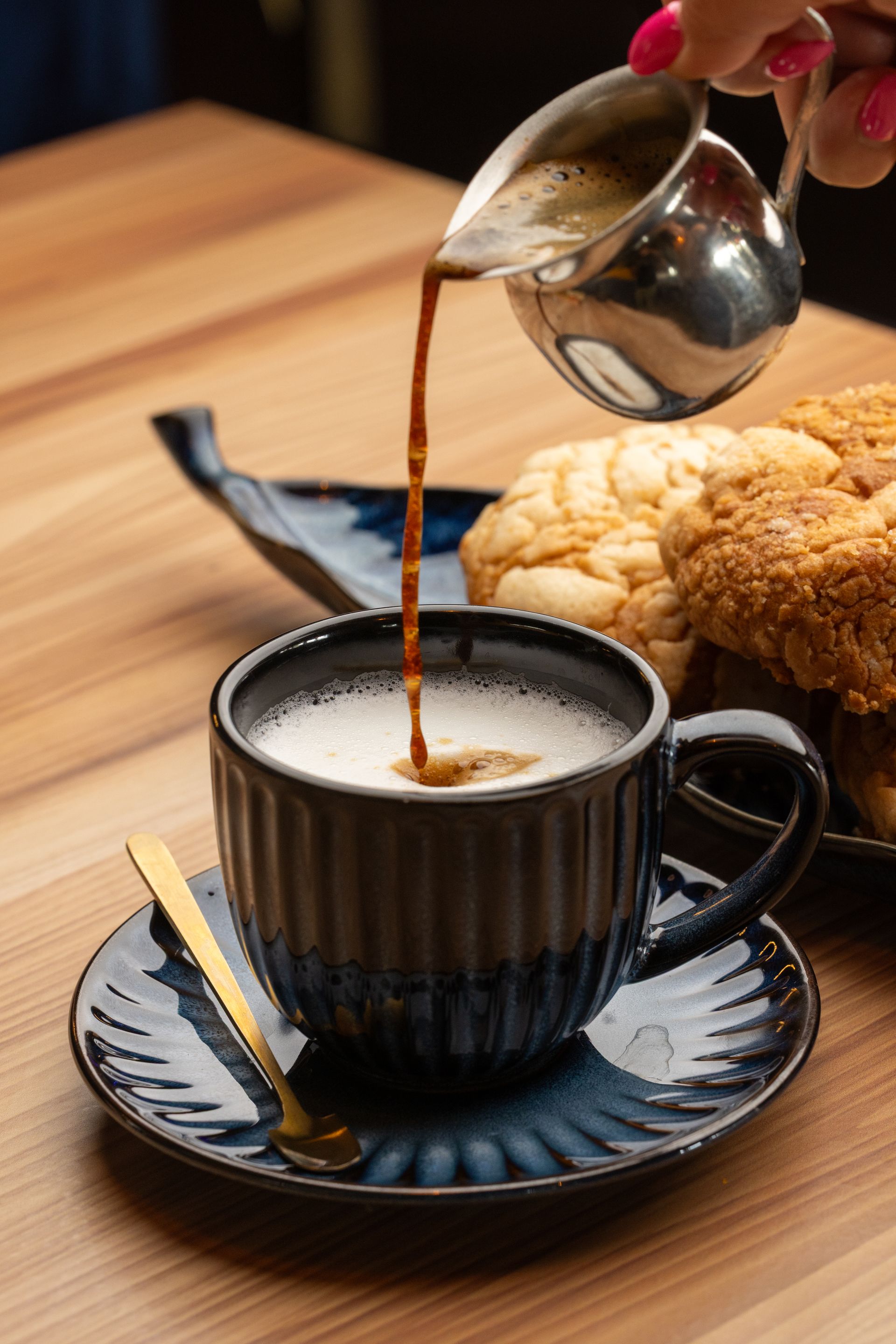 Coffee being poured into a black mug, with a saucer, spoon, and baked goods on a wooden table.