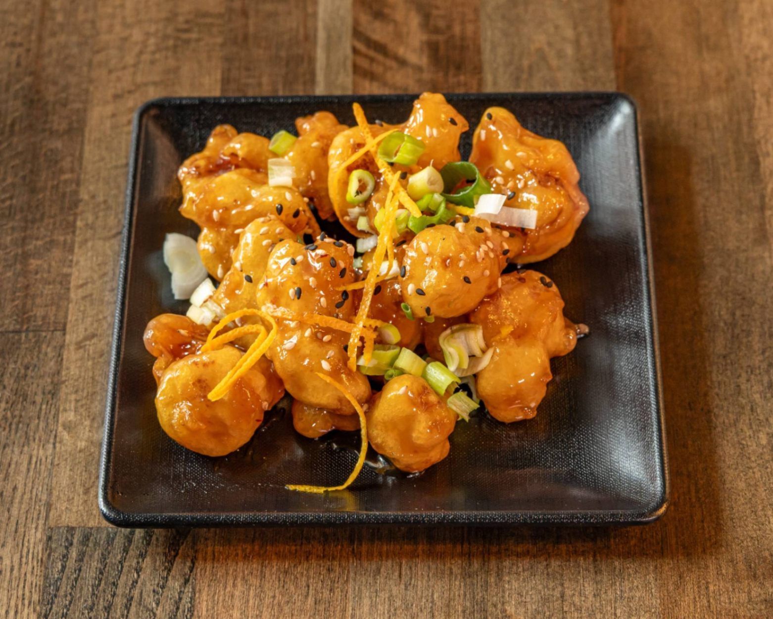 Orange-glazed fried bite-sized pieces topped with scallions and sesame on a black square plate