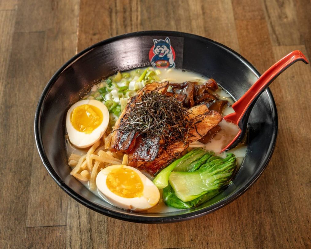 Black bowl of ramen with halved eggs, greens, sliced pork, and a red spoon on a wooden table