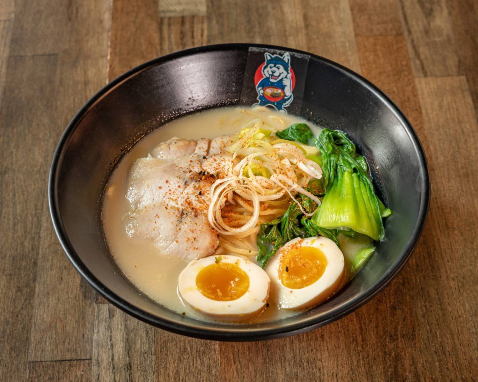Black bowl of creamy ramen with soft-boiled eggs, sliced pork, noodles, and green bok choy on a wooden table