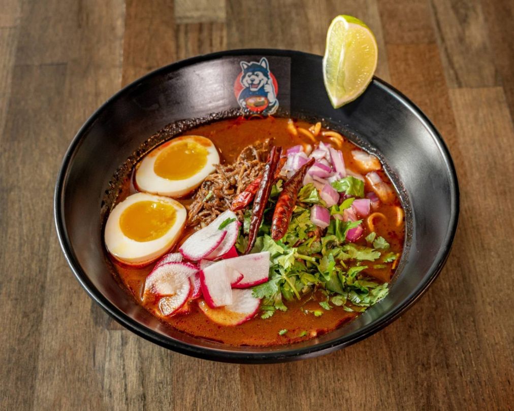 A bowl of spicy ramen with soft-boiled eggs, sliced pork, greens, and a lemon wedge on a wooden table.