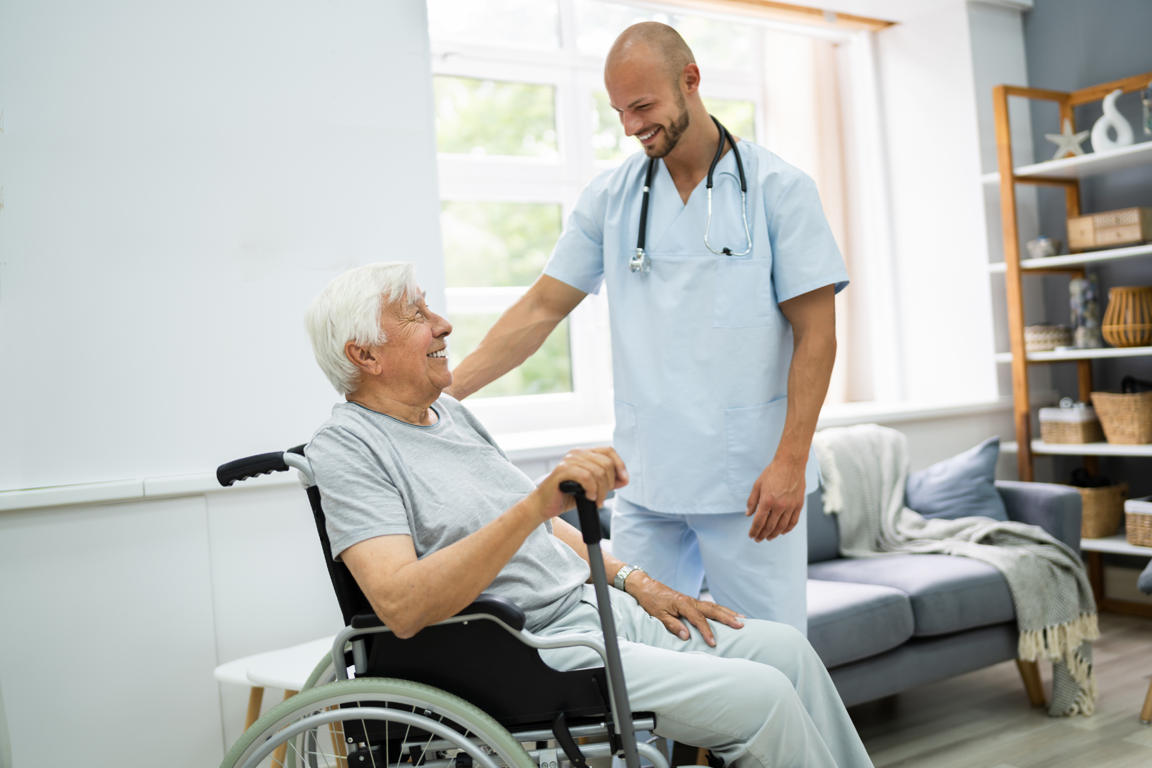 Male caregiver smiles at person in wheelchair, hand on shoulder, in home.