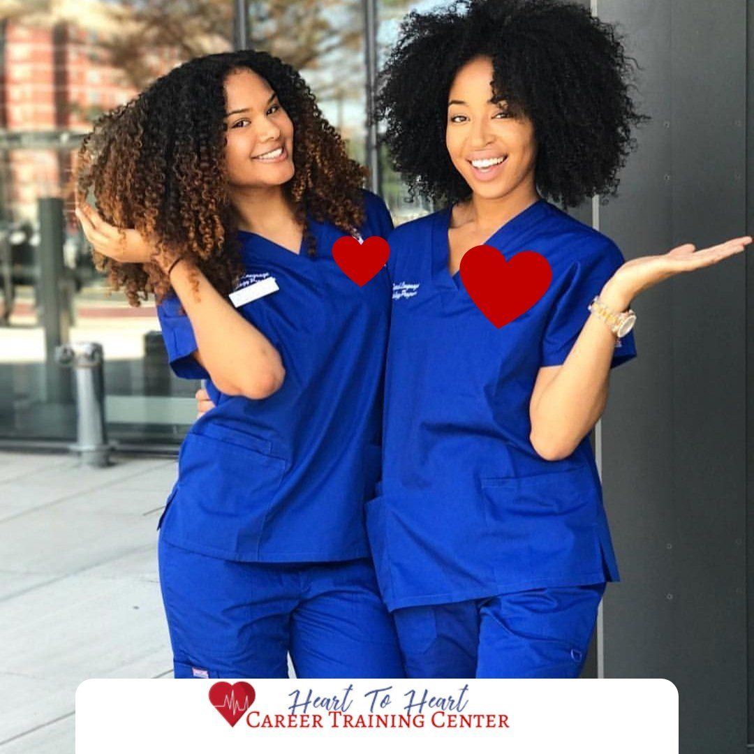 Two women in blue scrubs, smiling with hearts over their chests, posing outside.