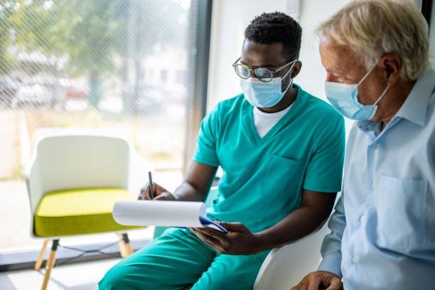 A medical professional in scrubs and a mask consults with a patient wearing a mask while holding a clipboard.