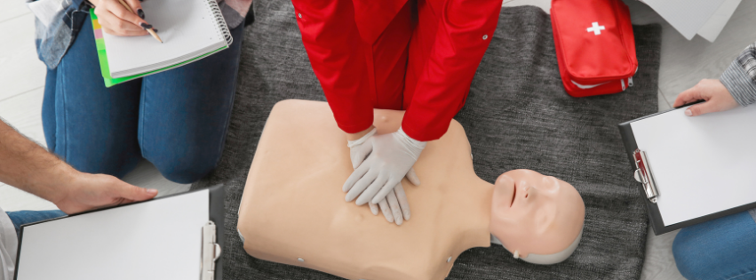 Person in red uniform performing CPR on a mannequin during a training class. Others are observing and taking notes.