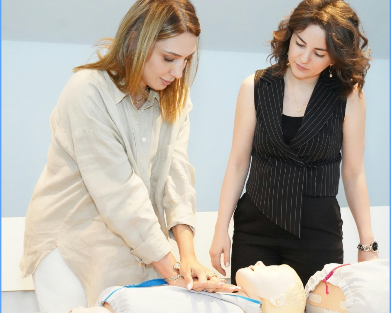 Two women demonstrate CPR on a training dummy. One woman presses on the chest, while the other watches.