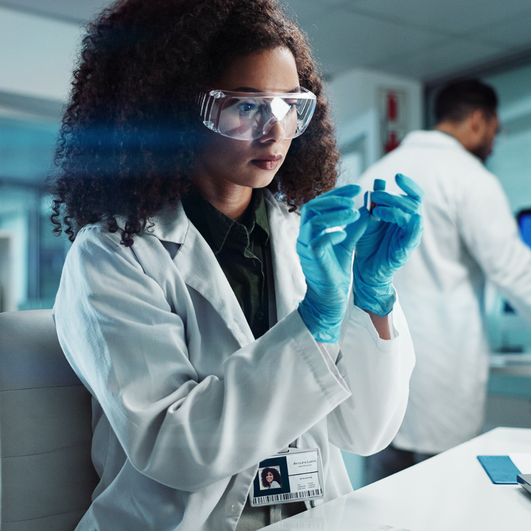 Woman in lab coat and safety glasses examining an object in a laboratory.