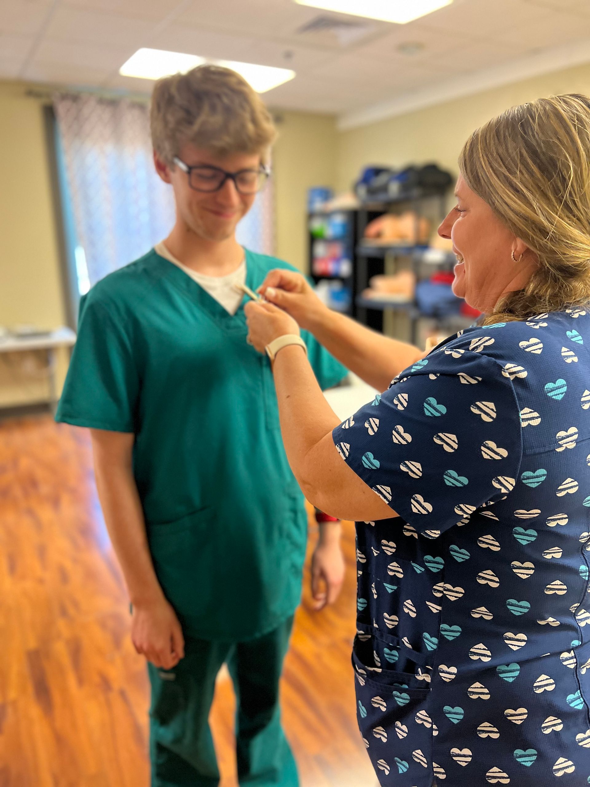 Woman pinning a badge on a person in teal scrubs. Interior setting, smiling, wooden floor.