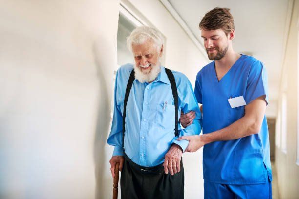 Elderly person using a cane, assisted by a person in blue scrubs, walking down a hallway.