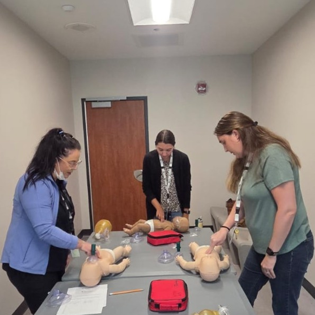 Three people practice infant CPR on training dummies in a classroom.