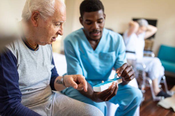 Caregiver showing an older person medication. Another person sits in the background. Indoor setting.