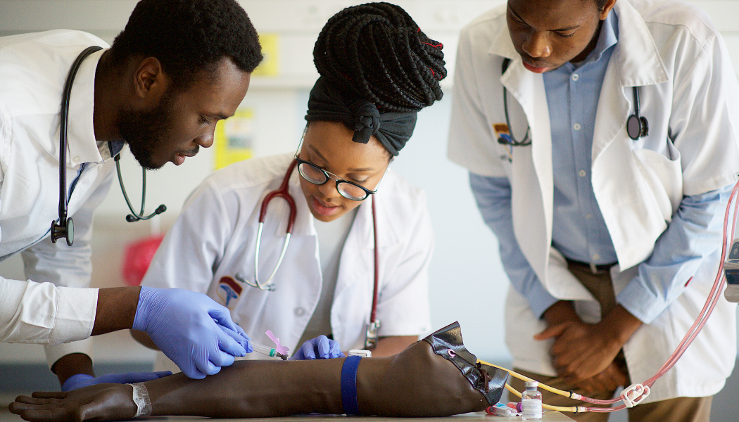 Three medical students practice injecting a mannequin arm in a training setting.
