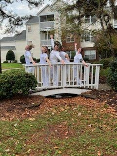 Five people in white outfits pose on a white bridge, arms raised, in front of a building.