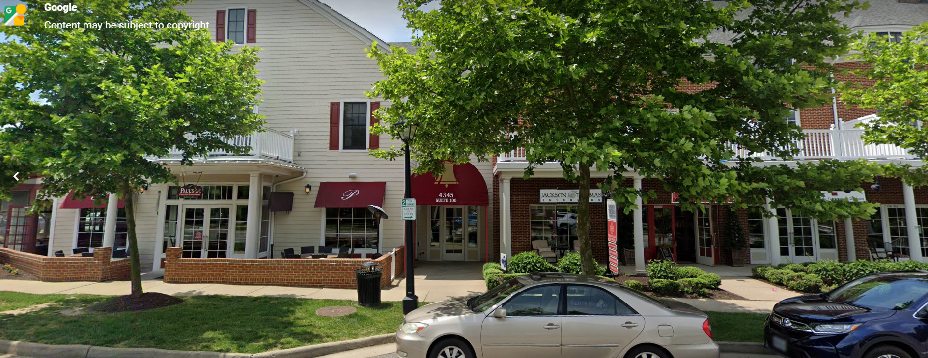 Shops with red awnings and trees on a street. A silver sedan and blue car parked.