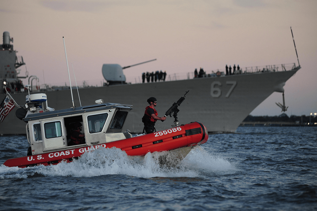 Coast Guard boat speeds past a large naval ship; both at sea.