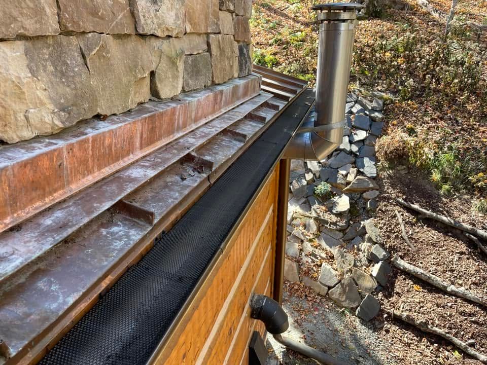 Rain gutter with leaf guard beneath a stone wall and metal chimney on a sloped roof.