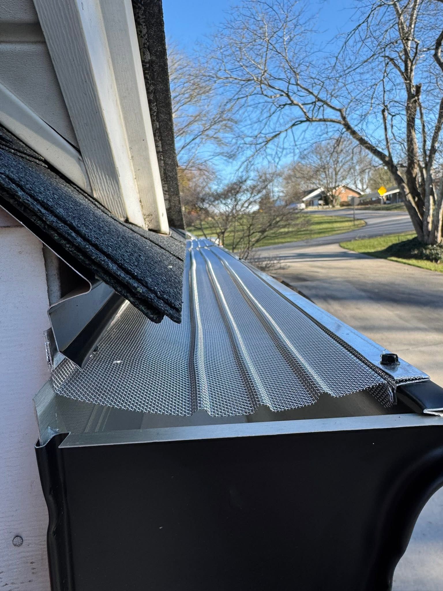Window awning with black support arms on a house exterior, seen beside a street and bare tree.