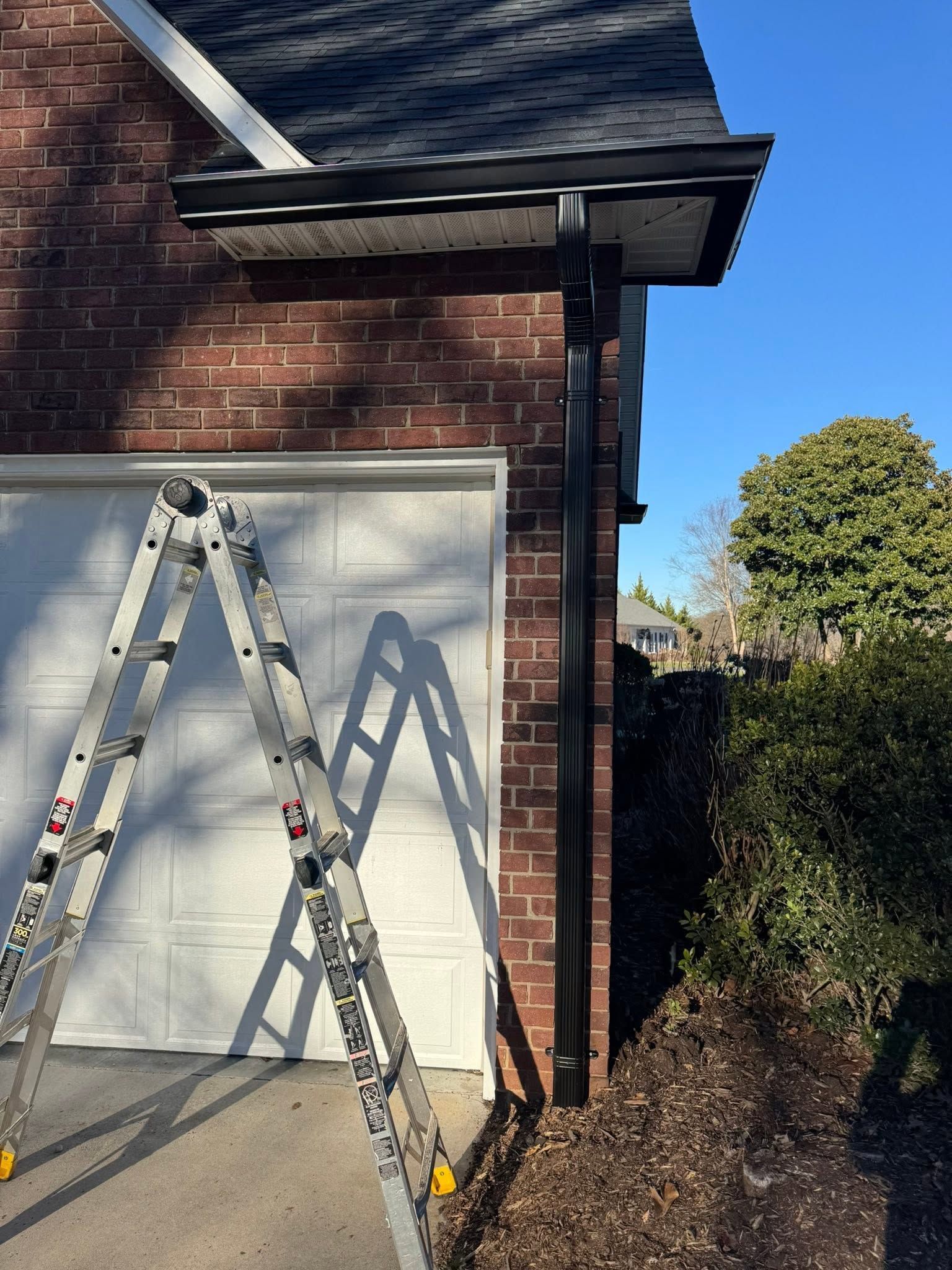 Two ladders leaning against a brick house beside a garage door, with a hedge on the right.