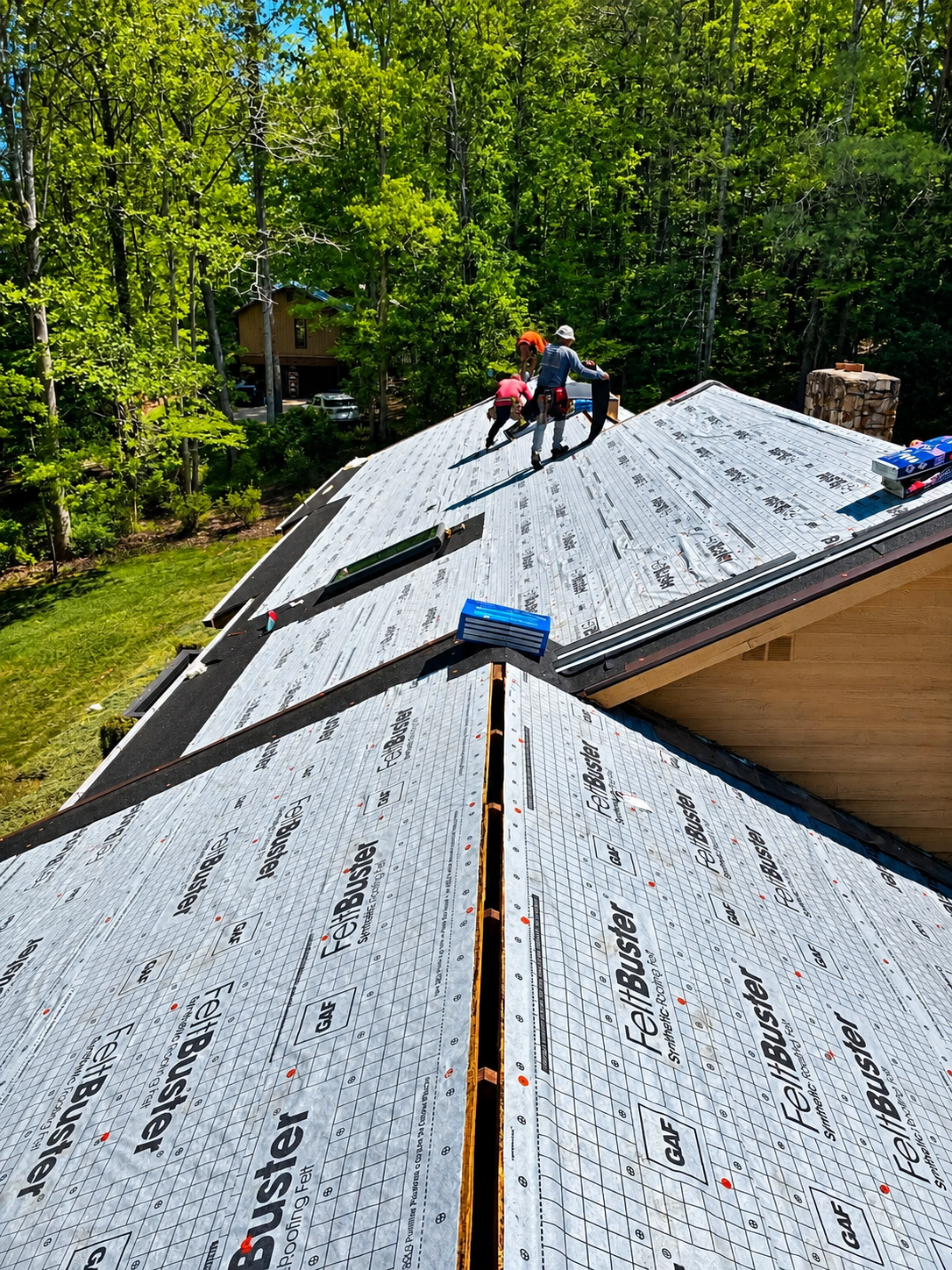 Workers on a roof installing roofing underlayment on a house surrounded by trees