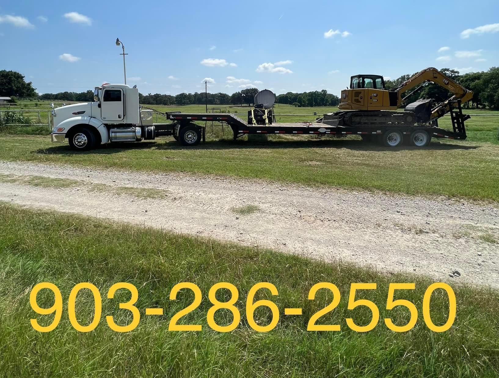 White truck hauling yellow excavator on a trailer on a gravel road, phone number in front.