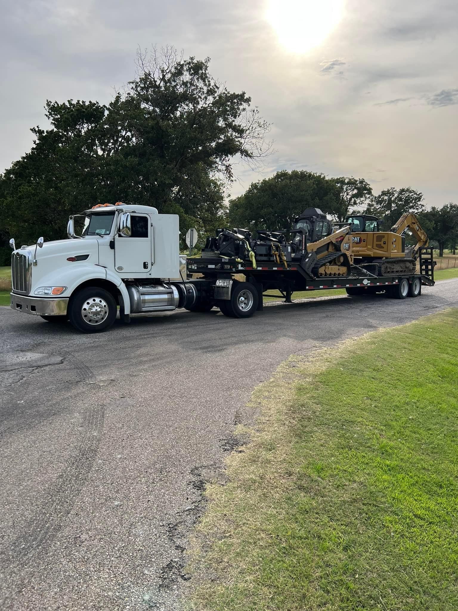 White semi-truck hauling construction equipment on a trailer, parked on gravel road. Green grass and trees in background.