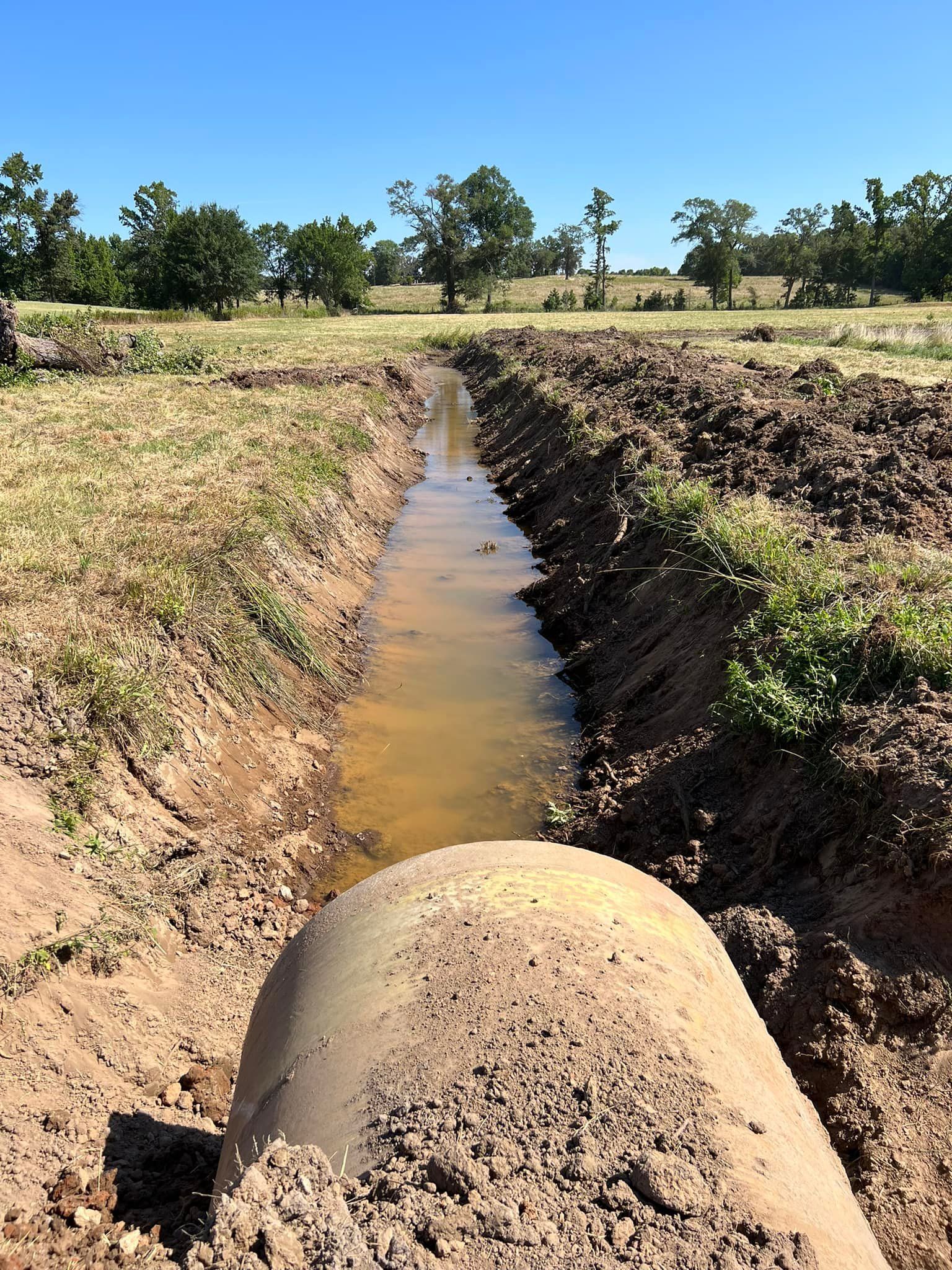 A muddy ditch filled with water, extending towards trees under a blue sky.