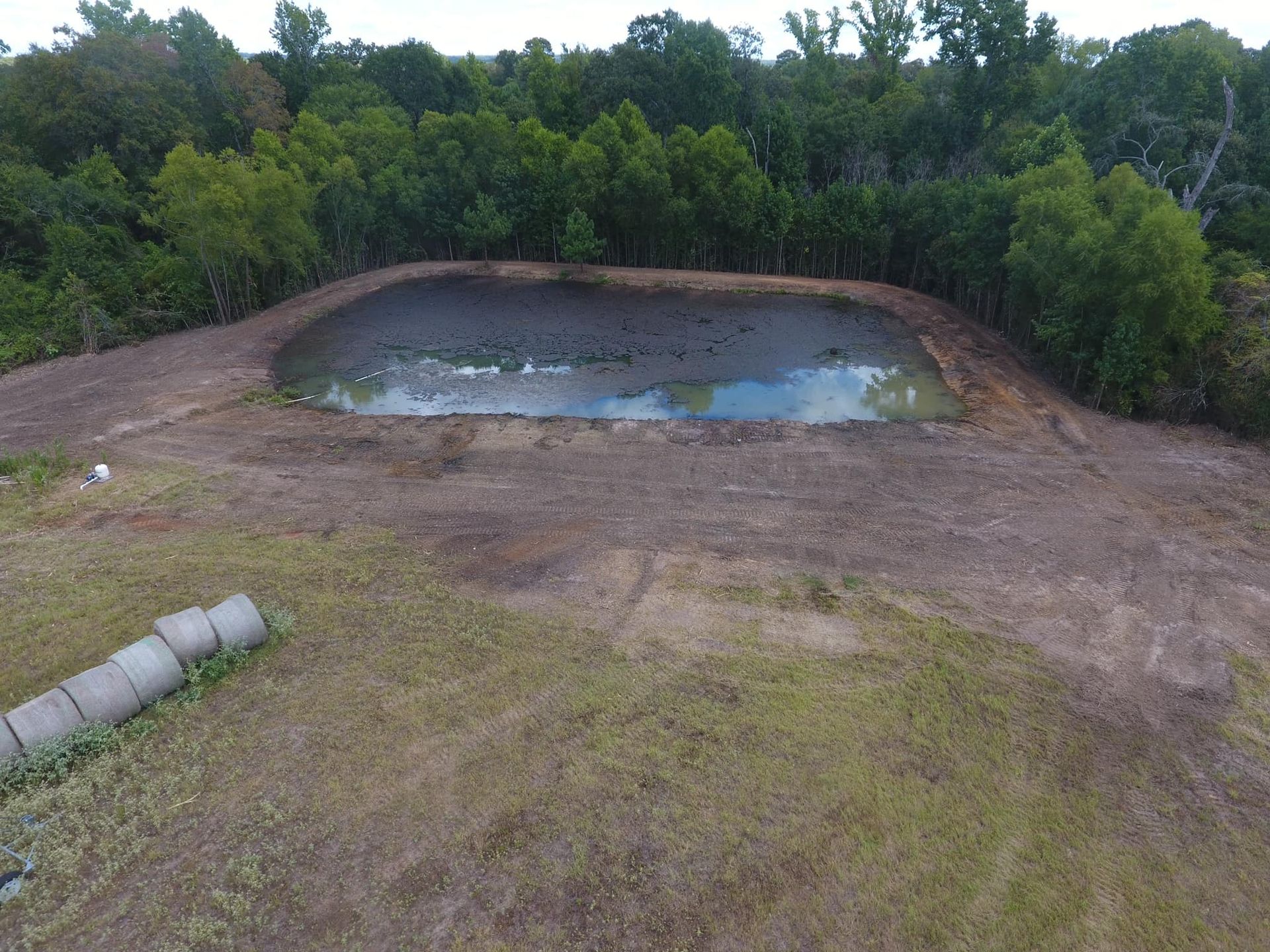 An aerial view of a pond surrounded by brown earth and trees. Hay bales lie in the foreground.