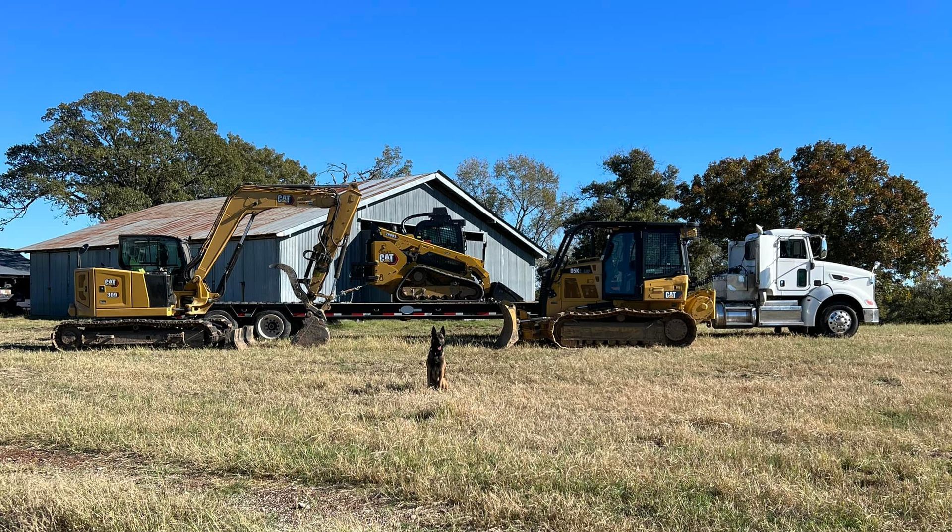 Excavator, bulldozer, and tow truck moving a small building across a grassy field under a blue sky.