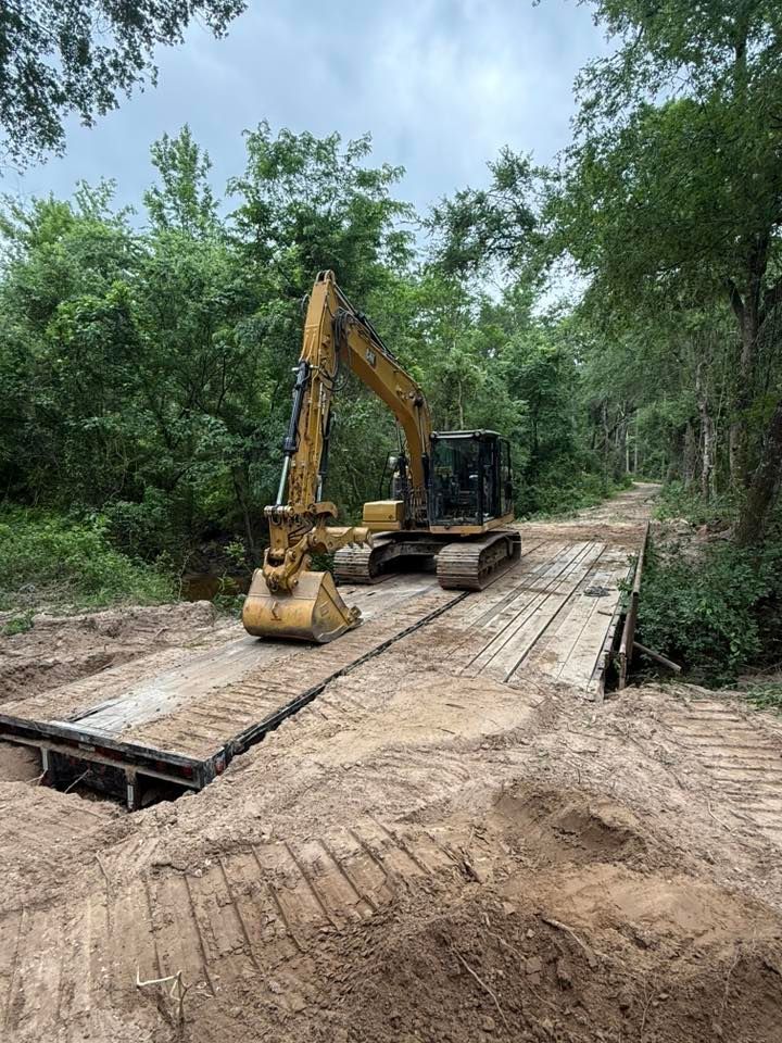 Yellow excavator on a wooden bridge in a wooded area, likely during a construction or repair project.