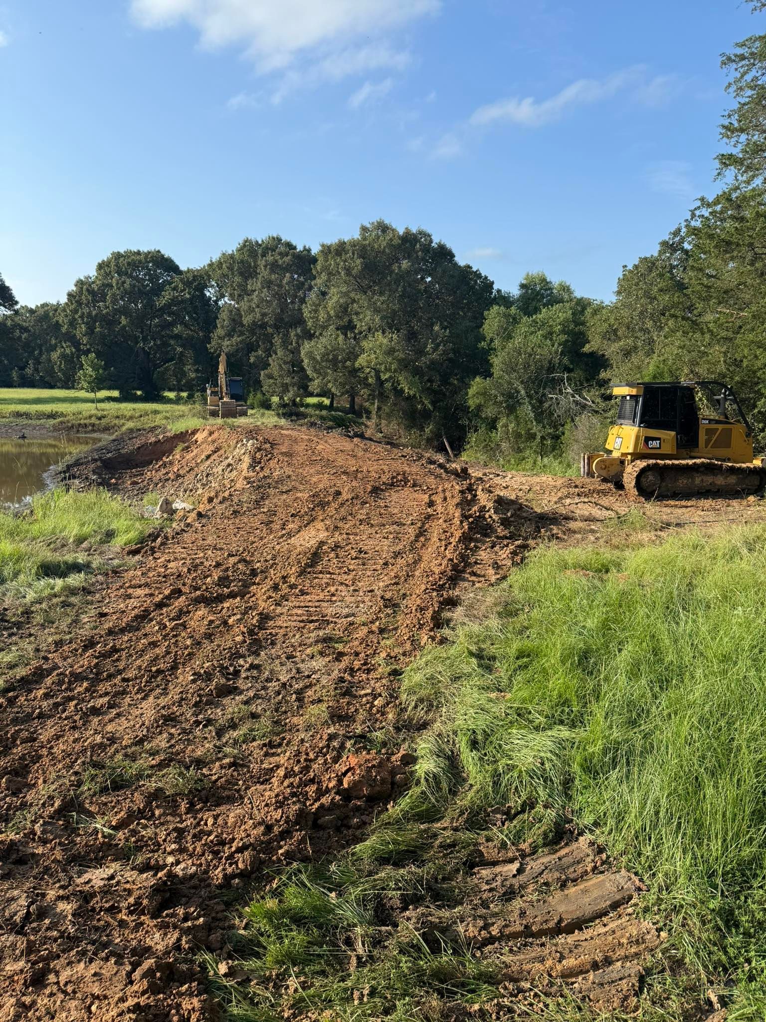 A small bulldozer on a dirt path near a pond, trees in the background.