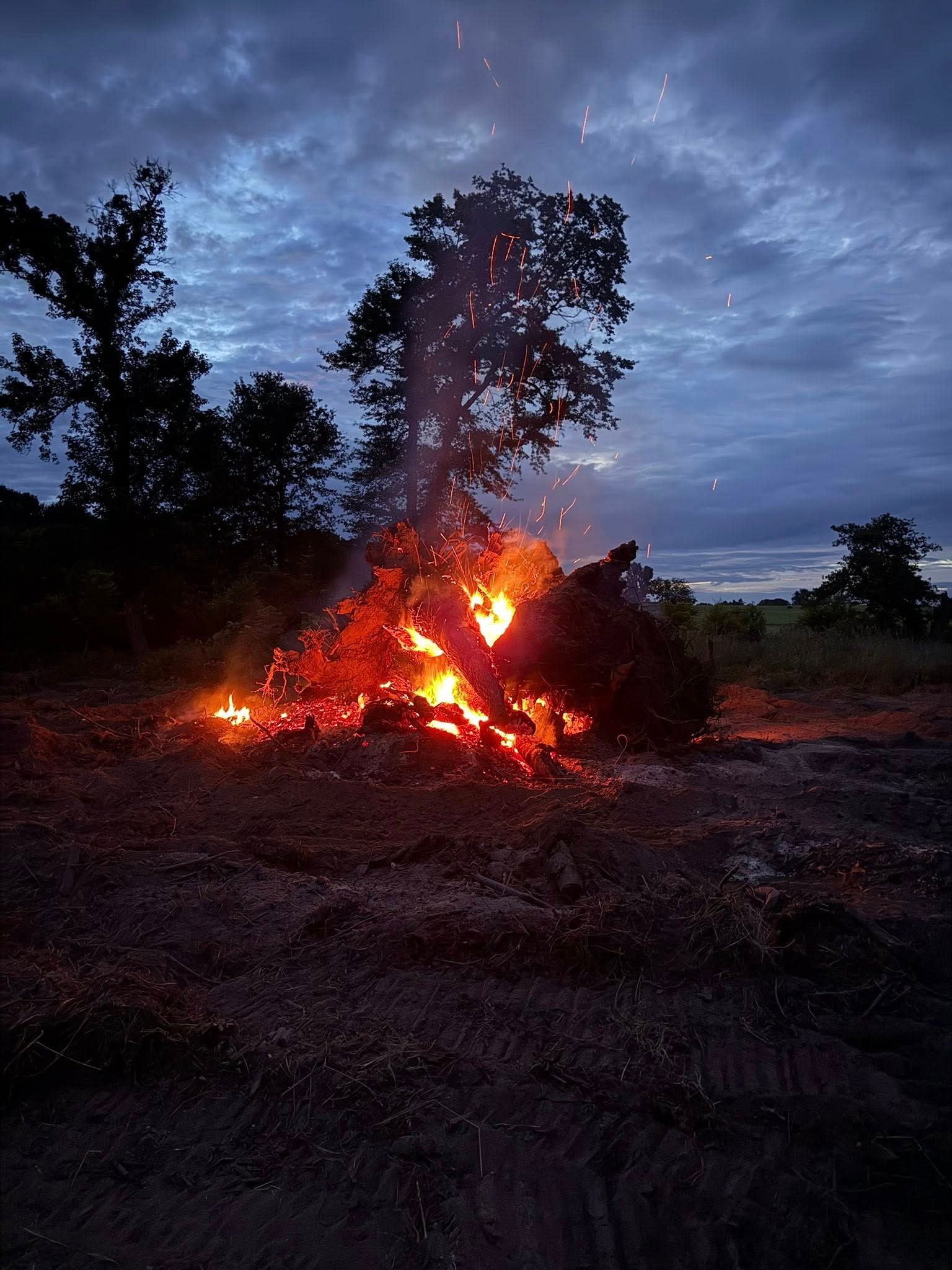 Burning bonfire at dusk with sparks rising, set against a dark cloudy sky.