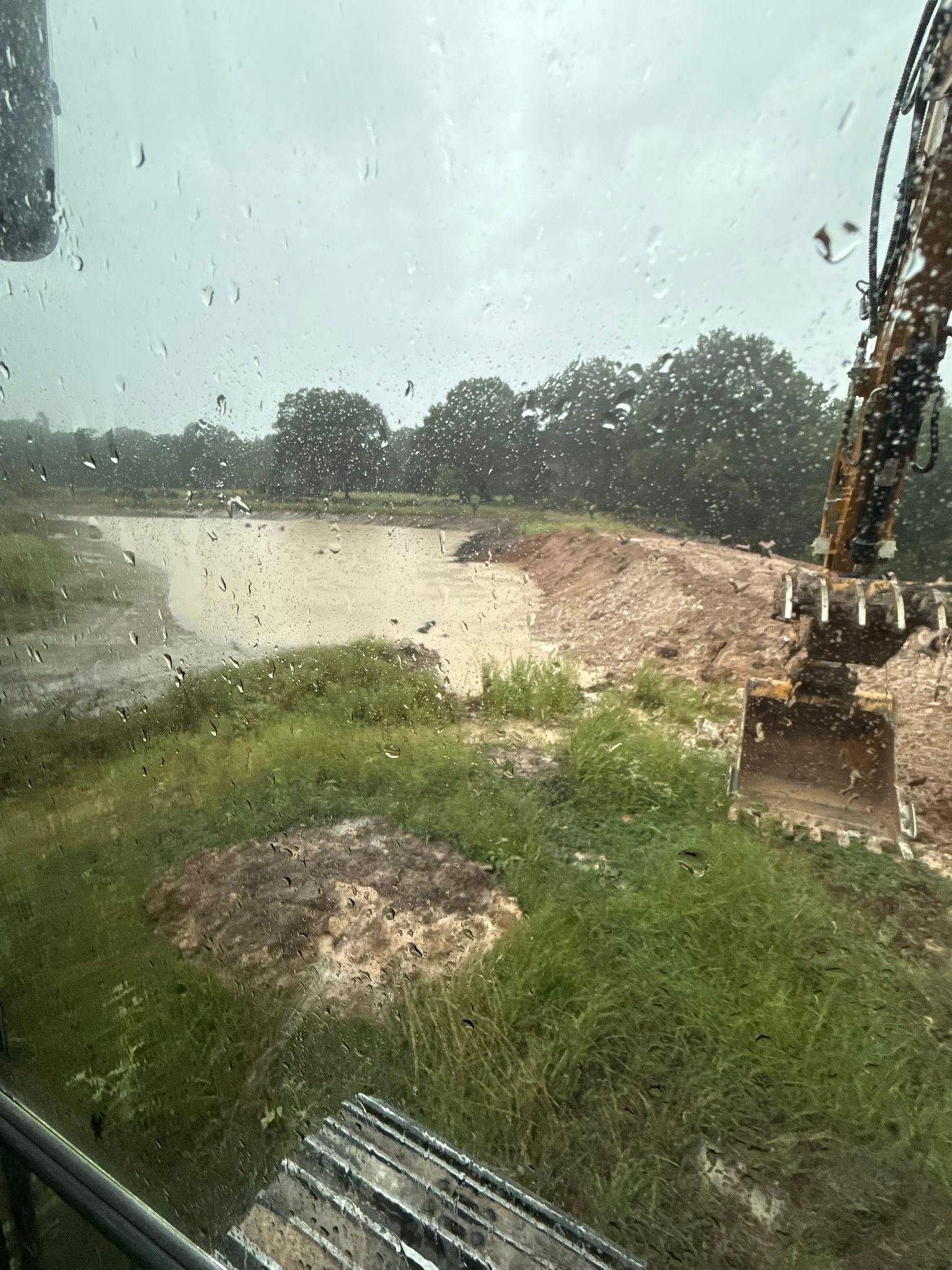 Rainy view from a construction vehicle: a muddy pond being dug, surrounded by trees and greenery.