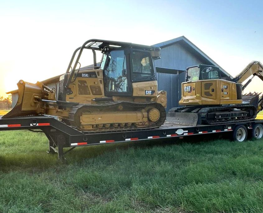 Two yellow Caterpillar construction machines on a trailer in a grassy field near a barn at sunset.