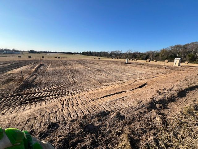 Dirt field with tractor tracks, hay bales, and trees under a clear blue sky.